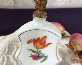 Op de afbeelding: Een witte porseleinen parfumfles met een gouden kroonvormige dop. De fles heeft een handgeschilderd bloemenmotief met een oranje tulp, roze en blauwe bloemen en groene bladeren.