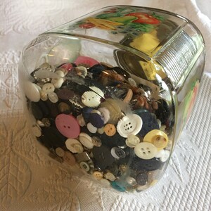 Puede incluir: Un tarro de cristal transparente lleno de una variedad de botones de diferentes colores, formas y tamaños. El tarro tiene forma rectangular con esquinas redondeadas y un diseño decorativo de frutas en la tapa. Los botones incluyen blanco, negro, rosa y azul.