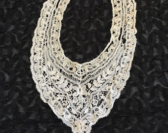 May include: An off-white, vintage lace collar with a scalloped edge and intricate floral and leaf patterns. The collar is U-shaped and appears to be handmade, with delicate detailing throughout. The lace is set against a dark, textured background.