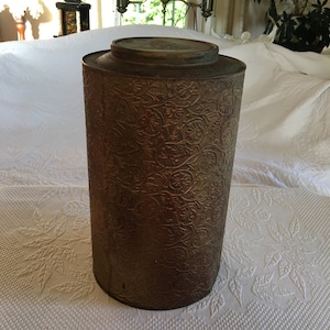 Puede incluir: Un recipiente de metal vintage, estampado con un diseño floral. El recipiente es de color marrón claro con un brillo dorado. Tiene una tapa y está en buen estado.