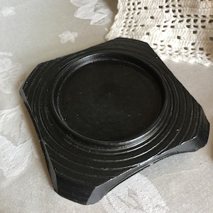 Puede incluir: Una base de madera negra con un centro circular y anillos concéntricos. La base es de forma cuadrada con bordes redondeados.