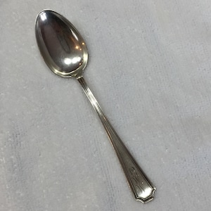 Puede incluir: Una cuchara plateada con un mango largo y delgado y un cuenco ovalado. El mango tiene un diseño decorativo cerca del extremo. La cuchara probablemente está hecha de acero inoxidable o plata y está diseñada para comer.