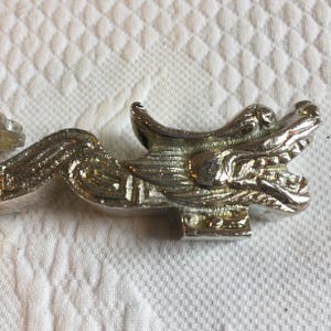 Könnte beinhalten: Ein silberfarbener Schubladengriff aus Metall mit einem detaillierten Drachenkopf-Design, offenem Maul und texturierten Schuppen. Der längliche Körper des Drachen erstreckt sich horizontal und zeigt ein metallisches Finish mit Reliefs.