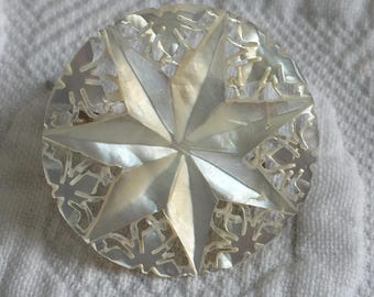 Puede incluir: Un botón redondo e iridiscente con un diseño de estrella. El botón presenta una estrella central de ocho puntas, rodeada por un borde decorativo calado. El botón está hecho de un material nacarado, que refleja la luz con un brillo sutil.