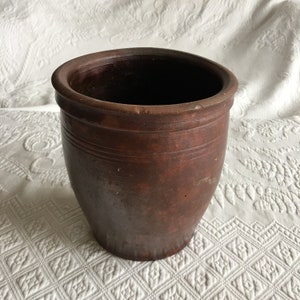 May include: A brown, cylindrical ceramic pot with a slightly flared rim. The pot has a textured surface and horizontal grooves around the middle. The interior is dark, and the pot appears to be empty. The pot is likely for decorative or functional use.