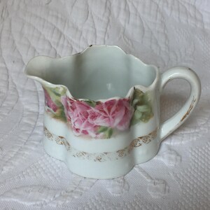 Puede incluir: Una jarra de crema de porcelana blanca con un borde festoneado y un asa. La jarra está decorada con rosas rosas y detalles dorados.