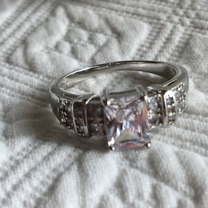 May include: A silver ring with a large, square-cut, clear gemstone surrounded by smaller clear gemstones.