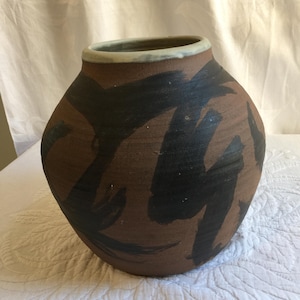 May include: A brown ceramic vase with a wide body and a narrow neck. The vase is decorated with bold black brushstrokes. The rim is a light beige colour. The vase has a rustic, handmade appearance.
