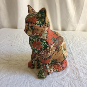 May include: A ceramic cat figurine covered in a patchwork of colorful floral fabric. The cat is sitting with its head turned to the side.