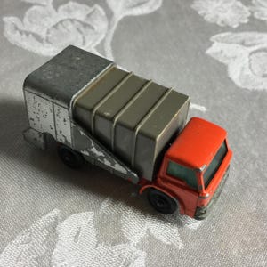 Puede incluir: Un camión de juguete vintage con cabina roja y caja gris. El camión tiene ruedas negras y carrocería plateada. El juguete muestra signos de desgaste, lo que sugiere que es un objeto de colección muy apreciado.