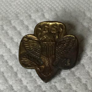 May include: A gold-colored Girl Scout pin featuring an eagle with outstretched wings and the letters "GS" above the eagle. The eagle is holding a shield with the American flag on it.