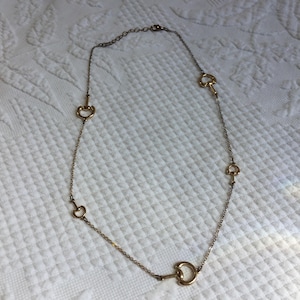 May include: A gold-toned necklace featuring a delicate chain with evenly spaced, decorative horse bit charms. The necklace is displayed on a white, textured surface, showcasing its elegant design and craftsmanship.