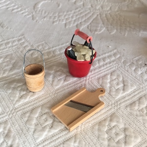 Puede incluir: Un conjunto de tres artículos de cocina en miniatura: un cubo rojo con asa lleno de cubitos de hielo blancos, un cubo de madera con asa de metal y una tabla de cortar de madera con una cuchilla de metal.