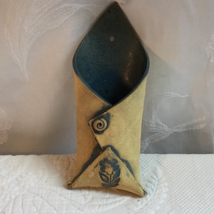 May include: A handmade ceramic wall pocket vase. The vase is a folded design with a tan exterior and a dark blue interior. A blue floral design is on the front, and a spiral design is on the side. The vase is approximately 10 inches tall.