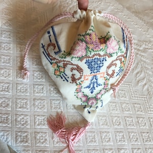 Puede incluir: Una bolsa de cordón blanco con un cordón y un borla de color rosa. La bolsa está bordada con un diseño floral en tonos de rosa, azul, verde y marrón.