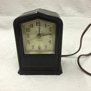 May include: A vintage black electric clock with a rectangular face and a decorative frame. The clock face is cream-colored with black numerals and hands. The brand name "Sessions" is printed above the center, and "Electric" is printed below. A brown cord extends from the back.