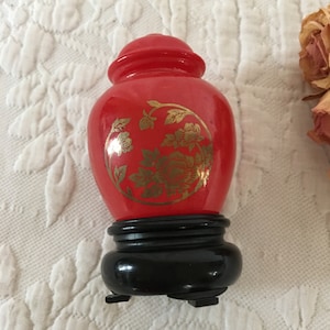 Op de afbeelding: Een rode keramische pot met een bloemenmotief. De pot heeft een zwarte houten voet met drie poten.