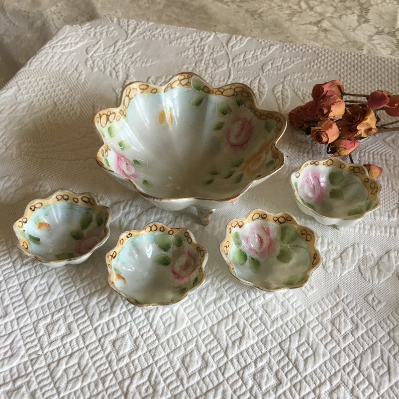 May include: A set of five white porcelain bowls with a scalloped edge and a floral design. The bowls are decorated with pink and yellow roses and green leaves. The bowls have a gold trim around the edge.