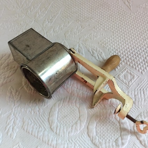 May include: An antique, hand-cranked food grater. The grater has a silver-colored metal drum and a white-painted frame with a wooden handle. The clamp-style base allows it to be secured to a countertop.