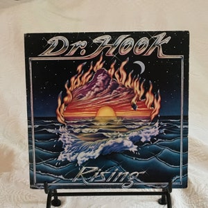 Puede incluir: Portada del álbum "Rising" de Dr. Hook. La obra de arte presenta una puesta de sol sobre olas del océano, con una montaña en llamas. El título del álbum y el nombre de la banda se muestran en una fuente estilizada. El fondo es azul oscuro con estrellas.