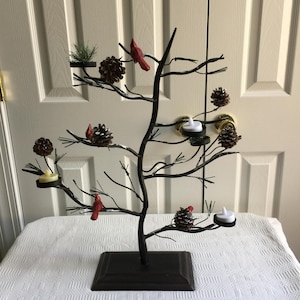 May include: A decorative metal tree with branches, pinecones, red cardinal figurines, and small candle holders. The tree is dark brown and sits on a square base. The candles are white. The background is a white door.
