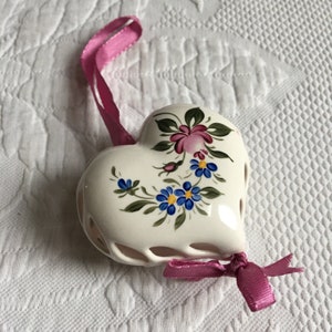 May include: A white ceramic heart ornament with a pink ribbon and a floral design featuring blue and pink flowers. The heart is openwork and has a delicate design.