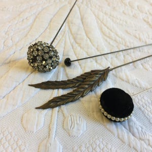 May include: Three vintage hat pins with decorative tops. One pin has a round, black velvet top with a pearl trim. Another pin has a round, clear rhinestone top. The third pin has a gold-toned feather design.