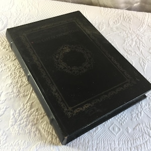 Könnte beinhalten: Ein schwarzes Hardcover-Buch mit goldener Schrift und einem dekorativen Rand. Der Titel "The East of the Morticians" ist auf dem Einband zusammen mit einem kreisförmigen Design zu sehen. Das Buch ist geschlossen und liegt auf einer weißen, strukturierten Oberfläche.