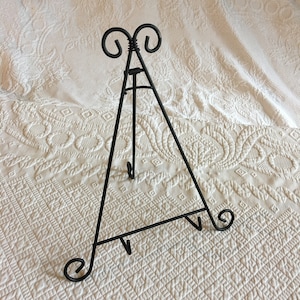 May include: Black metal easel stand with a decorative scroll design at the top. The stand is designed to hold a picture or other artwork.