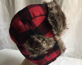 Wintermütze aus Polyester, Acryl und Viskose mit Kunstfellbesatz am Gesicht und Verzierungen auf rot-schwarz kariertem Fleece-Grundmuster. Mit Kinnverschluss.