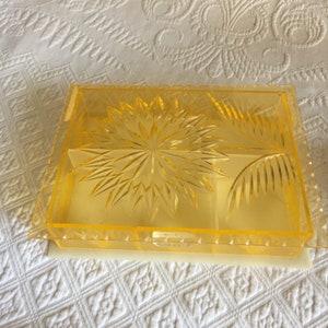 Puede incluir: Una caja de joyería de plástico transparente amarilla con un diseño floral. La caja tiene una tapa con bisagras y un compartimento para guardar joyas.