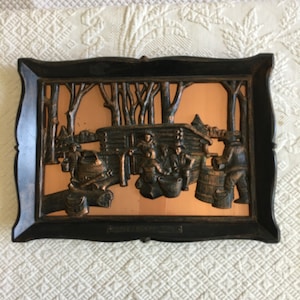 May include: A framed copper wall art piece depicting a scene of people making maple syrup in a forest. The scene is in relief and features a log cabin, trees, and people working with buckets and a cauldron.