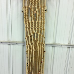 May include: A handmade macrame wall hanging in shades of beige and brown, suspended from a wooden dowel. The intricate woven design features geometric patterns and dangling fringe with decorative beads, creating a bohemian aesthetic.