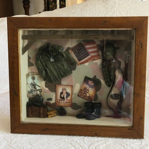 May include: A shadow box with a camouflage background featuring a green military jacket, a US flag, a military helmet, a rifle, a pair of boots, and several other military-themed items. The shadow box also includes two posters, one with the text "I Want You For U.S. Army" and the other with the text "Mustang Accessories".