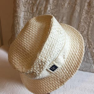 May include: A cream-colored woven straw hat with a slightly upturned brim. The hat features a dark blue square patch with a logo on the side. The hat is a classic accessory for sun protection and style.