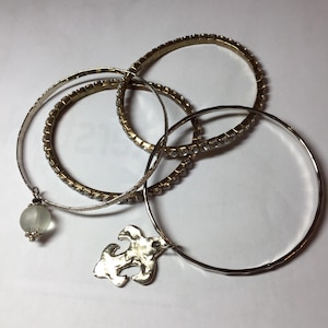 May include: An assortment of bracelets in gold and silver tones. One bracelet features a clear, frosted bead charm. Another has a silver charm with an abstract design. Several bracelets are adorned with small, sparkling rhinestones.