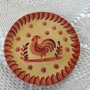 Puede incluir: Plato de cerámica decorativo con diseño de gallo. El plato tiene un centro amarillo con un gallo rojo, detalles florales rojos y un borde de hojas rojas. El plato es de forma redonda.