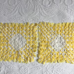 May include: Two square crocheted coasters featuring a textured, knotted pattern in bright yellow and white yarn. The design consists of alternating yellow and white loops arranged in a grid, creating a dense, tactile surface with fringed edges.