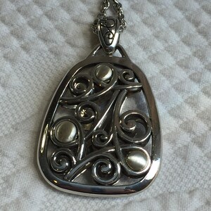 May include: A silver-toned metal pendant necklace featuring an openwork design with swirling scroll patterns and three smooth, circular, light-colored cabochon accents. The pendant is suspended from a matching silver-toned metal chain link.