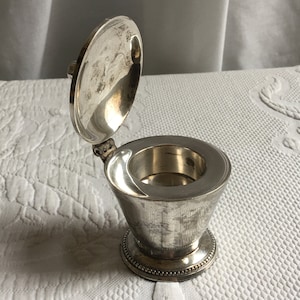 Puede incluir: Un recipiente plateado con tapa redonda y tapa con bisagras. La tapa está abierta y el recipiente está vacío.