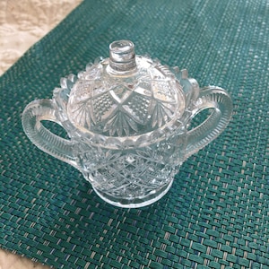 May include: A clear glass sugar bowl with a lid, featuring a cut-glass design. The bowl has two handles and a decorative lid with a pointed top.
