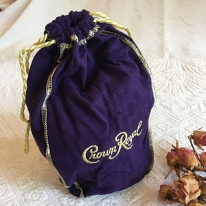 Könnte beinhalten: Eine violette Kordeltasche mit einem goldenen gestickten Crown Royal Logo. Die Tasche ist aus einem weichen, strukturierten Stoff.