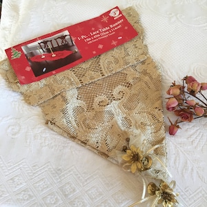 Op de afbeelding: Een beige kant tafelkleed met een delicaat bloemenpatroon. De tafelkleed is 33 cm x 114 cm en wordt geleverd met een decoratief ornament.