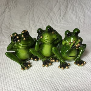 Peut inclure: Trois grenouilles en céramique verte, chacune avec un geste différent : l’une se couvrant les yeux, l’autre se couvrant les oreilles et la dernière se couvrant la bouche. Les grenouilles sont assises sur une surface blanche. La grenouille qui se couvre la bouche a le mot « Orlando » écrit sur son ventre.