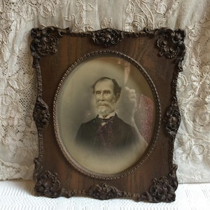 May include: An antique portrait of a man with a white beard in a dark suit, set within an ornate, dark brown wooden frame. The frame has decorative corner accents and a textured surface, adding to its vintage appeal.