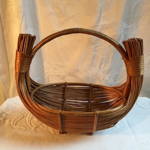 May include: A handcrafted brown wicker basket with a curved handle. The basket features a woven design with a rounded base and upright supports. The handle is securely attached to the basket's sides, providing a sturdy grip. The basket is suitable for carrying items.