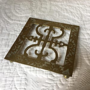 Puede incluir: Un salvamanteles de metal dorado con un diseño intrincado. El salvamanteles tiene forma cuadrada y está decorado con un patrón floral alrededor de los bordes. Tiene cuatro pequeñas patas en la parte inferior.