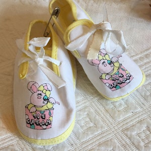 May include: A pair of white baby shoes with yellow trim and a bunny rabbit ice cream design. The shoes have white laces and are tied with a white ribbon.