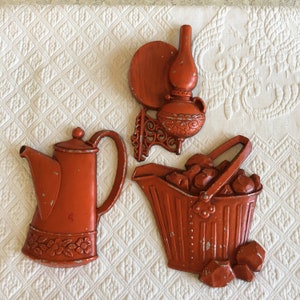 May include: Three vintage wall decor pieces in an orange color. The pieces are a teapot, a bucket with coal, and a lamp with a round shade.