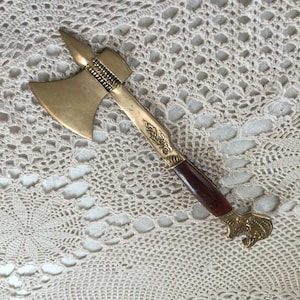May include: A decorative bottle opener shaped like a small axe. The opener features a brass-colored axe head and handle with a dark brown wooden grip. The handle has ornate detailing and a decorative end.
